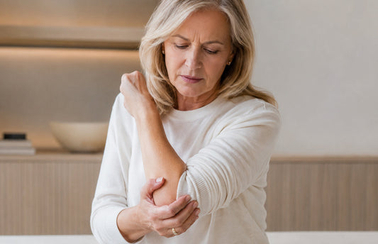 Mulher adulta madura segurando o cotovelo com expressão de desconforto em ambiente interno claro, representando atenção à perda de massa óssea, saúde dos ossos e avaliação da ingestão de cálcio.